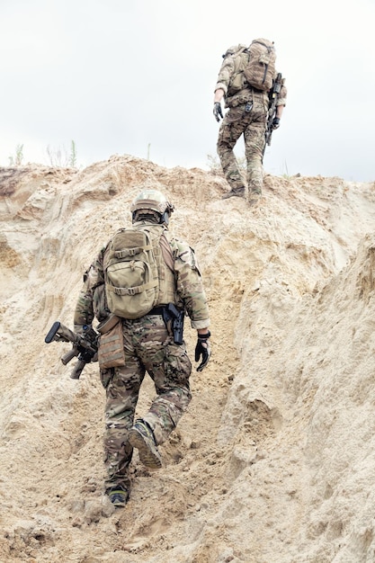 Zwei moderne Infanterie-, Spezialeinsatzkräfte-Soldaten, militärische Geheimdienstspezialisten, bewaffnet mit Dienstgewehren in schützender Tarnuniform mit Rucksack auf dem Rücken, die auf Sanddüne klettern