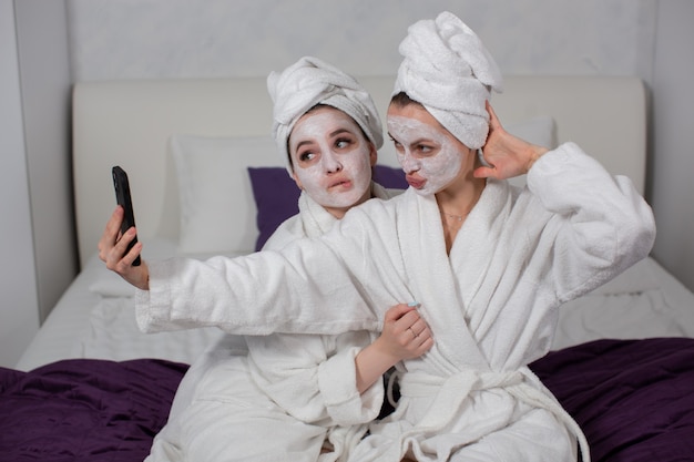 Zwei moderne Mädchen machen Spa-Behandlungen zu Hause für Gesicht und Körper und haben Spaß dabei, Selfies auf ihrem Telefon zu machen, hochwertiges Foto