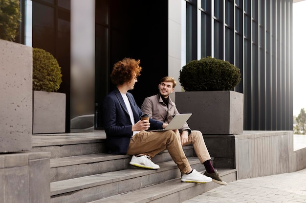 Zwei moderne, stylische Hipster, die auf der Treppe sitzen und mit einem Laptop Kaffee trinken Programmierer Konzept des erfolgreichen Geschäfts mit Remote-Arbeitspausen