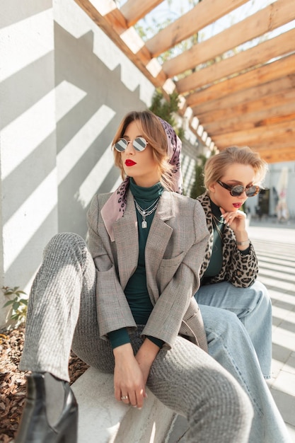 Zwei modisch schöne junge Mädchen mit Retro-Sonnenbrille in stylischem grauen eleganten Anzug und Leopardenjacke Blue Jeans sitzen und posieren an sonnigen Tagen auf dem Ochsen Trendige Hipster-Frauen in der Stadt