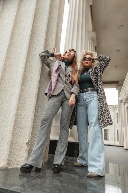 Zwei modische hübsche Models Mädchen mit cooler Sonnenbrille in trendiger Herbstkleidung mit Jeans-Leopardenmantel, grauem Anzug und Schuhen stehen in der Nähe von Vintage-Säulen auf der Straße