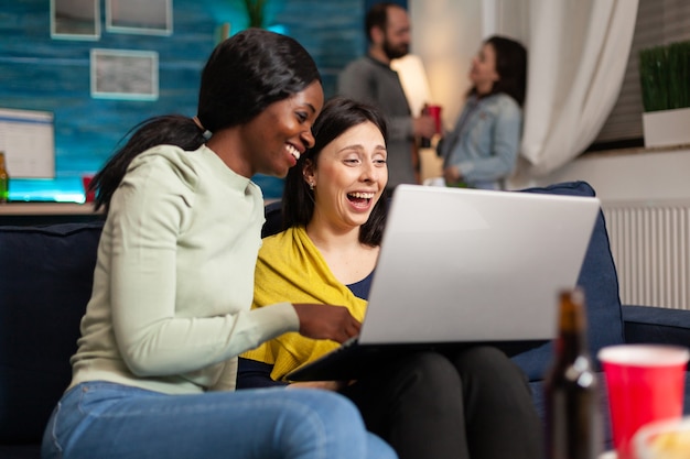 Zwei multiethnische Frauen sitzen auf der Couch und schauen sich Comedy-Serien auf Laptop-Computern an, die während der Nachtparty zusammen hängen. Im Hintergrund jubeln Freunde Bierflaschen zu, reden und haben Spaß.