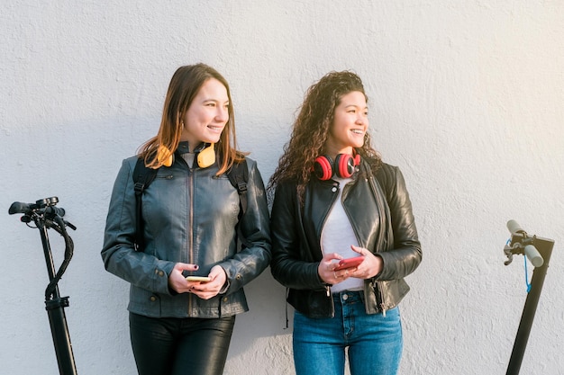 Zwei multiethnische Freundinnen auf Elektrorollern mit Smartphone im Freien