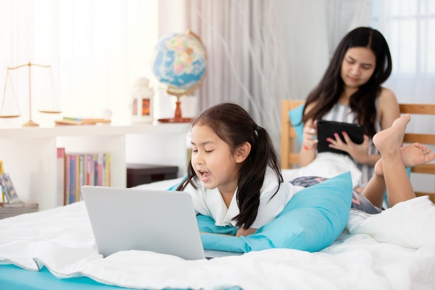 Zwei nette asiatische Kindermädchen, die Tablette und Laptop auf Bett verwenden.