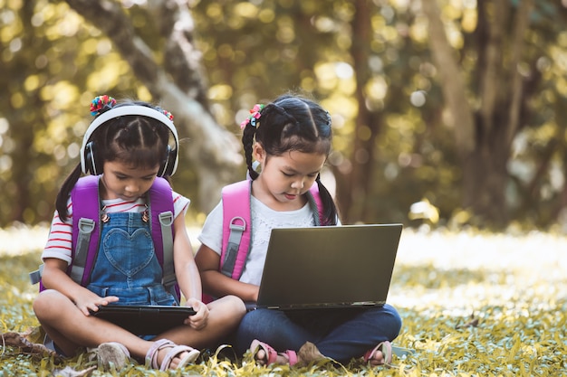 Zwei nette asiatische Kindermädchen, die zusammen Tablette und Laptop im Park verwenden