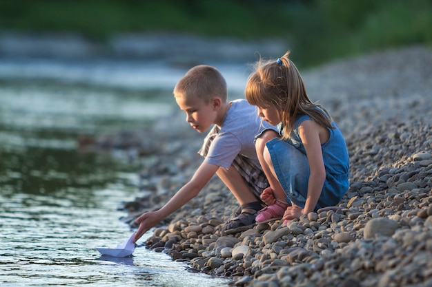 Zwei nette blonde Kinder, Junge und Mädchen auf der Flussbank, die Weißbuchboote des Wassers einsendet. Freuden und Spiele des glücklichen Kindheits- und Freientätigkeitskonzeptes.
