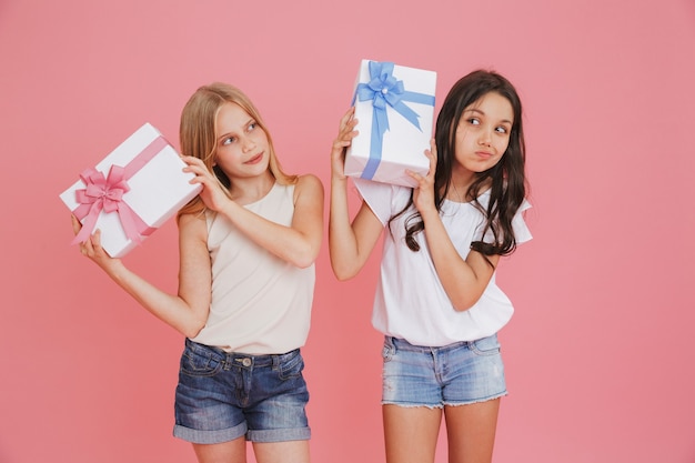 Zwei neugierige interessierte mädchen 8-10 in der freizeitkleidung, die geschenkboxen mit bunten schleifen hält und schüttelt, lokalisiert über rosa hintergrund