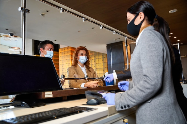 Zwei Personen in medizinischen Masken stehen beim Check-in hinter einem Glasschild in einer Hotelhalle
