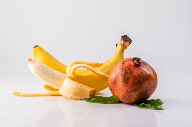 Zwei reife Bananen, eine geschält und eine Granatapfelfrucht mit Blättern auf weißem Grund.