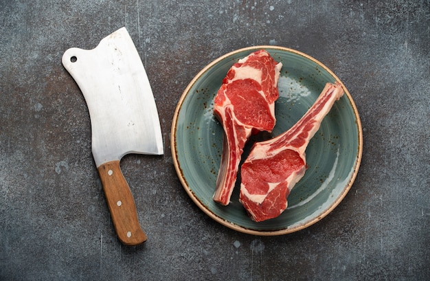 Zwei rohe marmorierte Tomahawk-Fleischsteaks auf Teller mit Fleischerbeil, Kräutern und Gewürzen, konkreter Hintergrund. Premium Beefsteak auf Knochenzubereitung, Draufsicht, Steakhaus oder Metzgereikonzept