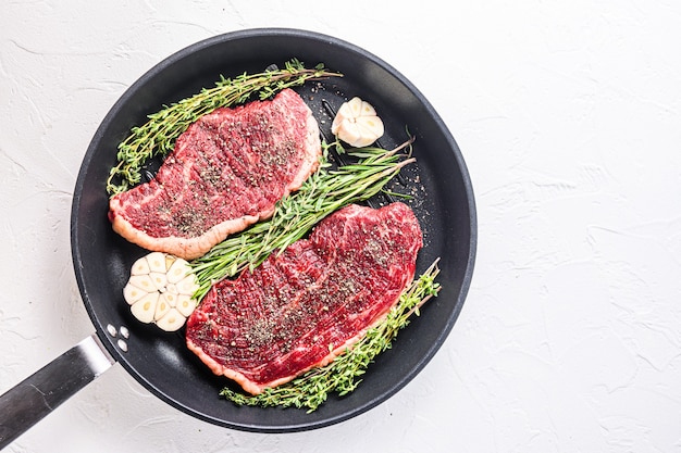 Zwei rohe Picanha-Bio-Rindfleischsteaks bereit zum Grillen auf Pfanne mit Kräutern und Knoblauch. Auf weißem strukturiertem Hintergrund Draufsicht mit Platz für Text.