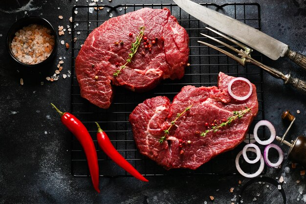 Zwei rohe Rindersteaks mit Gewürzen, Zwiebeln und Chili auf dunklem Schiefer oder Beton.