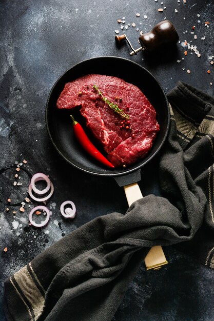 Zwei rohe Rindersteaks mit Gewürzen, Zwiebeln und Chili auf dunklem Schiefer oder Beton.