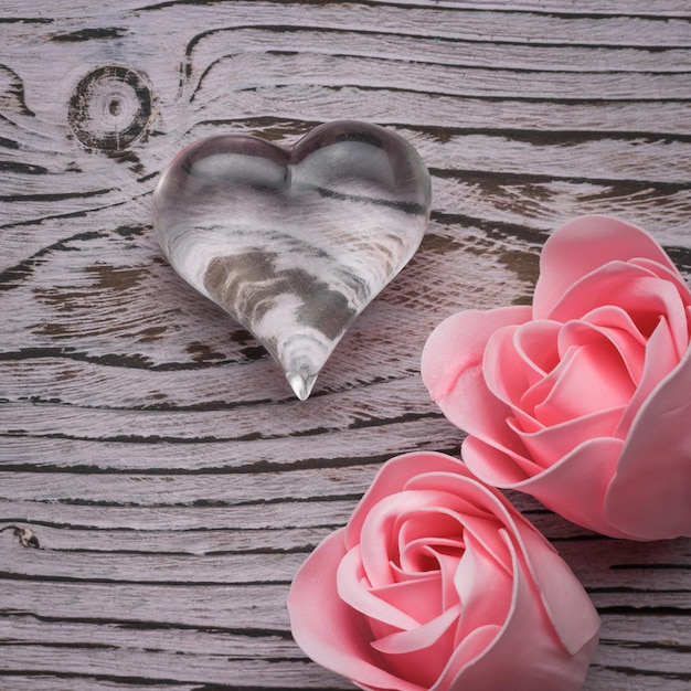 Zwei Rosenknospen und ein Glasherz auf einem hölzernen Hintergrund. Das Konzept der Liebe und des Valentinstags. Flach liegen.