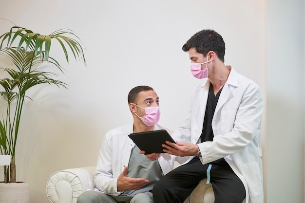 Zwei Ärzte im Wartezimmer mit einer Maske kommentieren die Ergebnisse mit einer Tablette in der Hand. Gynäkologische, zahnärztliche oder ästhetische Klinik. Medizinisches Konzept.