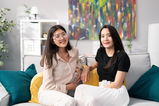 Zwei schöne asiatische Frauen, die sich auf der Couch im neuen Haus entspannen, junge Mädchen plaudern und reden über lustige Dinge