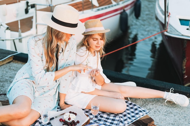 Foto zwei schöne blonde freundinnen, mutter und tochter in weiß und strohhüten, die auf der yacht am pier picknicken