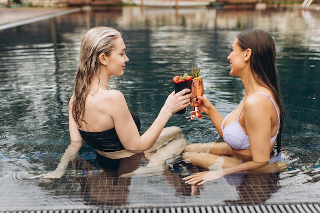 Zwei schöne Frauen, brünett und blond, unterhalten sich, trinken Cocktails und entspannen sich im Sommer am Pool