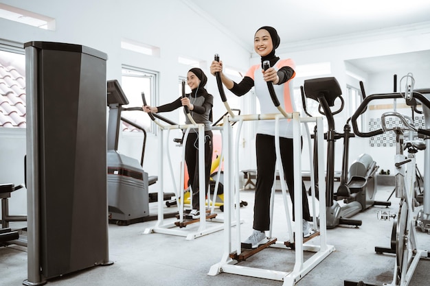 Zwei schöne Frauen mit Hijab trainieren im Fitnessstudio mit einer Freundin auf einem statischen Crosstrainer