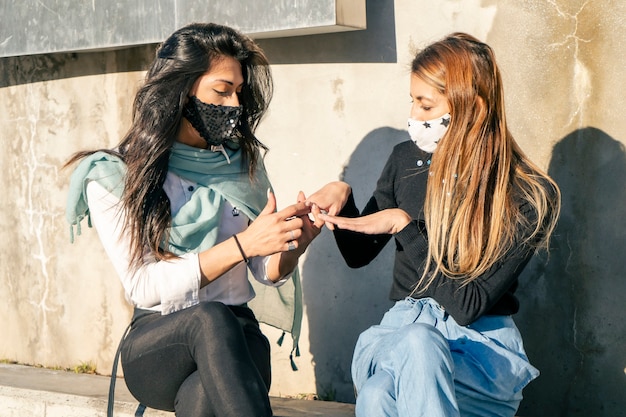 Zwei schöne Frauen mit Mundbedeckungen aus der COVID-Pandemie sitzen beim Reden und zeigen ihre Nägel. Freundschafts- und Sicherheitskonzept.