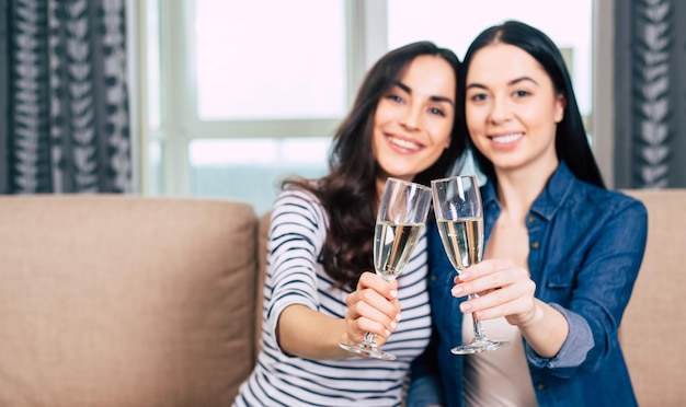 Zwei schöne Freundinnen in Freizeitkleidung sitzen auf der Couch und unterhalten sich und trinken Champagner