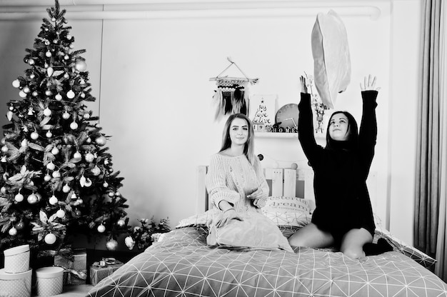 Zwei schöne Freundinnen tragen einen warmen Pullover und Beinwärmer (Gamaschen) spielen mit Kissen auf dem Bett gegen den Neujahrsbaum mit Weihnachtsdekoration.