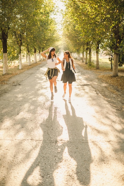 Zwei schöne, fröhliche junge Frauen mit Rucksäcken auf dem Rücken amüsieren sich beim Spaziergang entlang der sonnigen Allee.