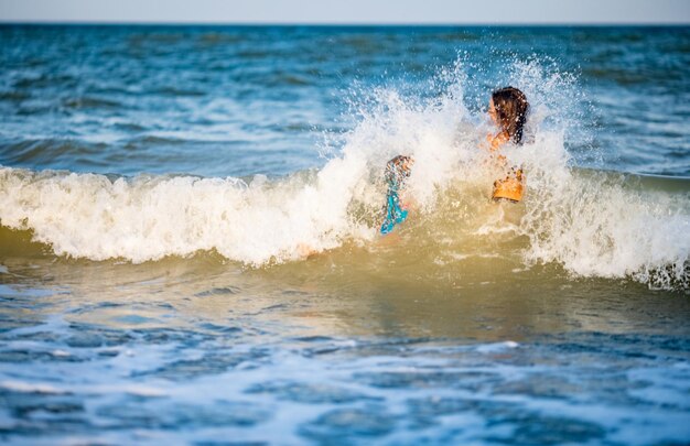 Zwei schöne, gebräunte, langhaarige, fröhliche Schwestern in hellen Sommerbadeanzügen kriechen und springen an einem warmen, sonnigen Urlaubsabend in blau schäumenden Meereswellen auf einer sandigen Untiefe mit kleinen Muscheln