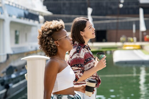 Zwei schöne gemischtrassige Frauen, die mit einem Kaffee, Booten und Yachten im Hintergrund durch den Hafen laufen