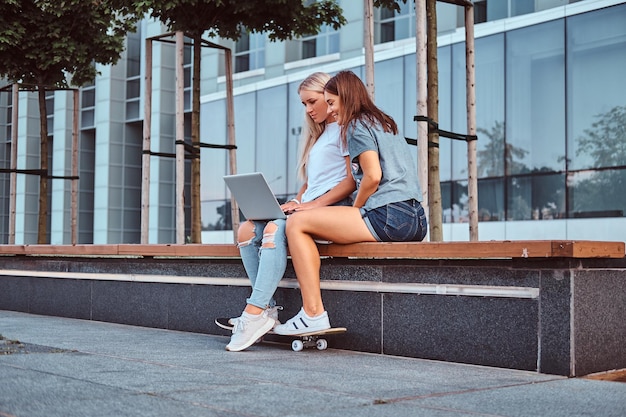 Zwei schöne Hipster-Mädchen, die einen Laptop benutzen, während sie auf einer Bank auf dem Hintergrund des Wolkenkratzers sitzen.