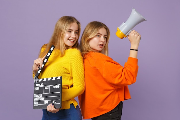 Zwei schöne junge blonde Zwillingsschwestern Mädchen halten klassische schwarze Filmklappe, schreien auf Megaphon isoliert auf violettblauer Wand. Menschen-Familien-Lifestyle-Konzept. .