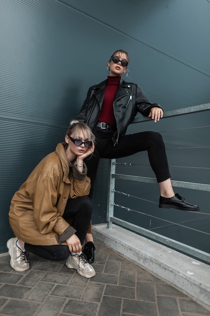 Zwei schöne junge Frauen in trendiger Denim-Kleidung mit einer Lederjacke in Sonnenbrille, die im Freien in der Nähe einer Metallwand posiert