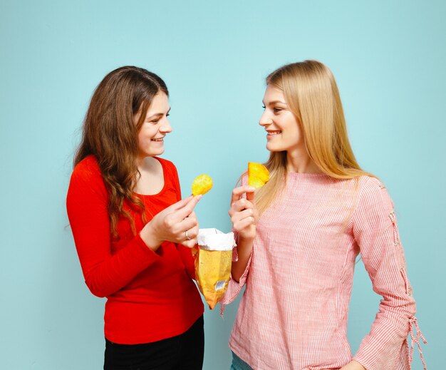Zwei schöne junge Mädchen, die Chips essen und sind auf einem blauen Hintergrund nett