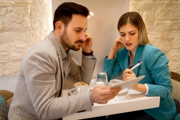 Zwei schöne junge nachdenkliche Geschäftsleute sitzen im Café und arbeiten an einem digitalen Tablet.