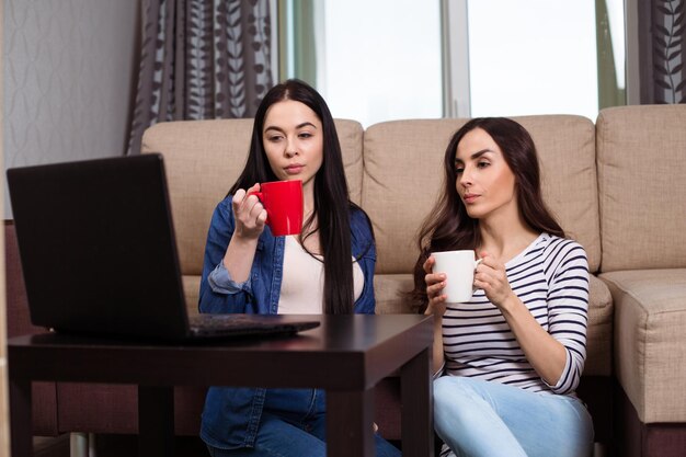 Zwei schöne junge und lächelnde Freundinnen in Freizeitkleidung sehen zu Hause auf dem Sofa fern oder Filme und trinken Kaffee oder Tee