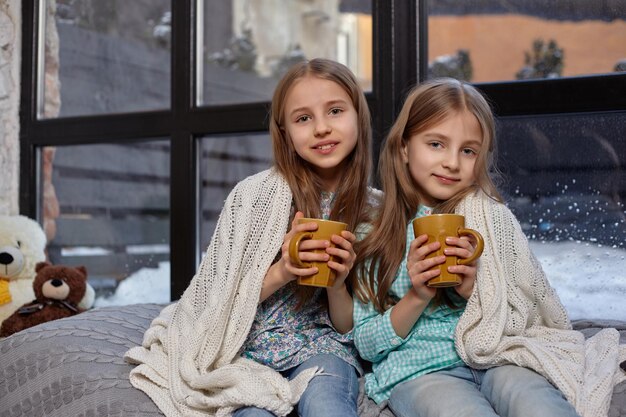 Zwei schöne kaukasische kleine Schwestern, die auf der Fensterbank sitzen, bedeckt von einer Decke. Sie erstarrten und versuchten sich aufzuwärmen und tranken köstlichen warmen Tee