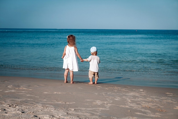 Zwei schöne kleine Kinder gehen in einer Tagesuhr an der Küste entlang und halten ihre Hände. Sie sind in weißer Kleidung und Hut