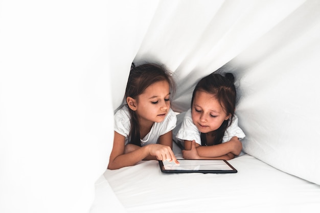 Zwei schöne kleine Schwestern, die im Bett liegen und auf den Bildschirm eines Tablets schauen, intelligente Kinder, die intelligente Technologie verwenden