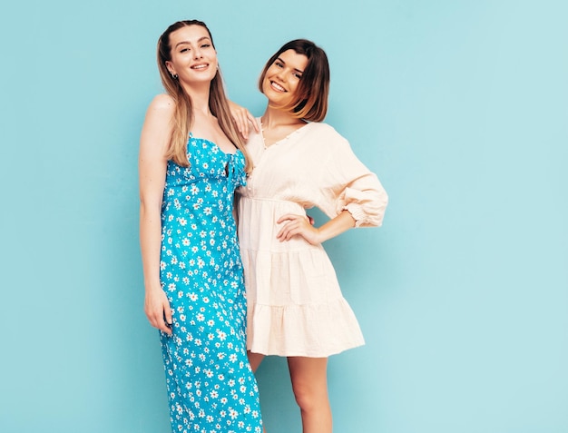 Zwei schöne lächelnde Hipster-Frauen in Sommerkleidung posieren in einem Studio in der Nähe einer blauen Wand.