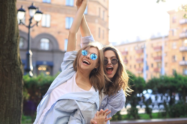 Zwei schöne lächelnde Mädchen in trendiger Sommerkleidung posieren auf Straßenhintergrund. Models haben Spaß und umarmen sich.