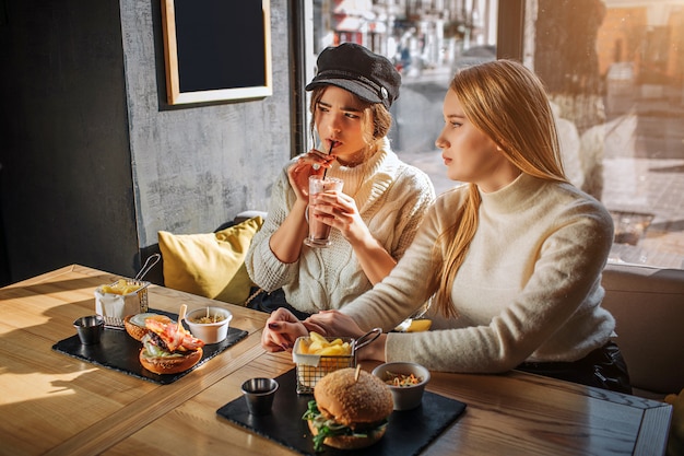 Zwei schöne Teenager sitzen am Tisch im Café und schauen nach links. Erster Cocktail trinken. Zweitens sitzen neben Freund. Sie sind aat Fenster. Draußen scheint die Sonne.