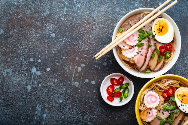 Zwei Schüsseln japanische Nudelsuppe Ramen mit Fleischbrühe, geschnittenem Schweinefleisch, Narutomaki, Ei mit Eigelb auf rustikalem Steinhintergrund. Traditionelles japanisches Gericht, Ansicht von oben, Nahaufnahme, Konzept. Platz für Text