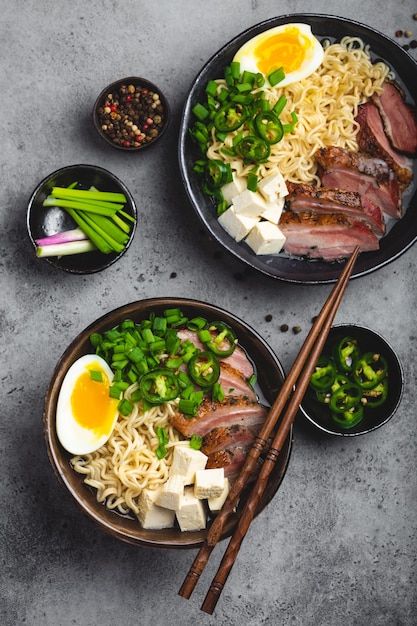 Zwei Schüsseln leckere asiatische Nudelsuppe Ramen mit Fleischbrühe, Tofu, Schweinefleisch, Ei mit Eigelb auf grauem rustikalem Betonhintergrund, Nahaufnahme, Draufsicht. Heiße leckere japanische Ramen-Suppe zum Abendessen nach asiatischer Art