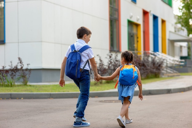 Zwei Schulkinder ein kleines Mädchen und ein Junge in einem weißen Hemd mit Rucksäcken machen sich auf den Schulweg
