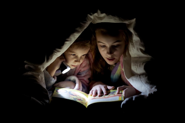 Zwei Schwestermädchen lesen im Dunkeln mit einer Taschenlampe ein Buch unter einer Decke
