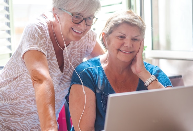 Zwei Schwestern, die das gleiche Laptoplächeln verwenden. Attraktive reife Frauen, die Kopfhörer teilen