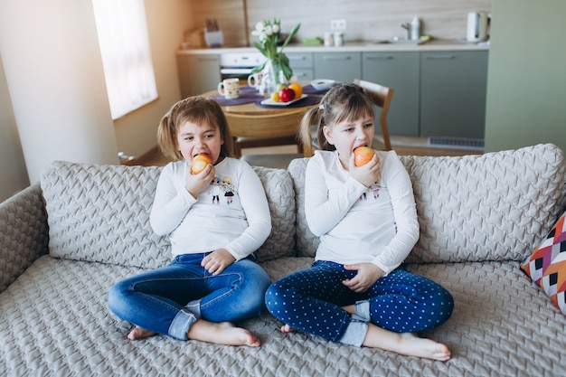 Zwei Schwestern in der ähnlichen Kleidung, die zusammen auf Sofa sitzt und Apfel isst