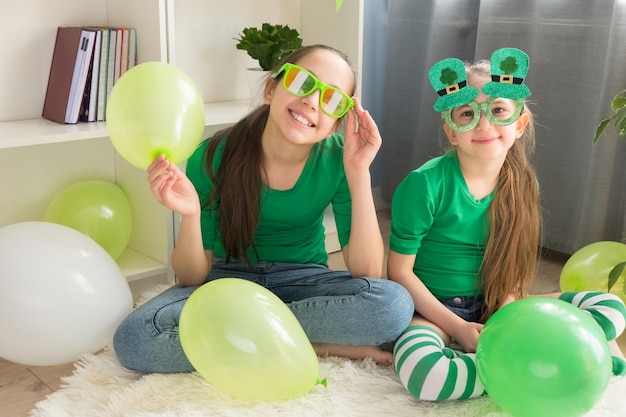 Zwei Schwestern in lustigen grünen Gläsern halten Luftballons und haben Spaß zu Hause beim Feiern des St. Patricks Day