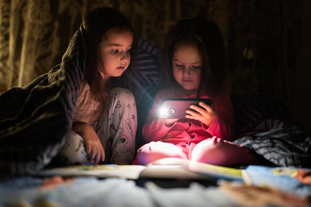 Zwei Schwestern schauen sich nachts zu Hause Filme oder Zeichentrickfilme auf dem Smartphone zusammen mit einer Decke im Bett an