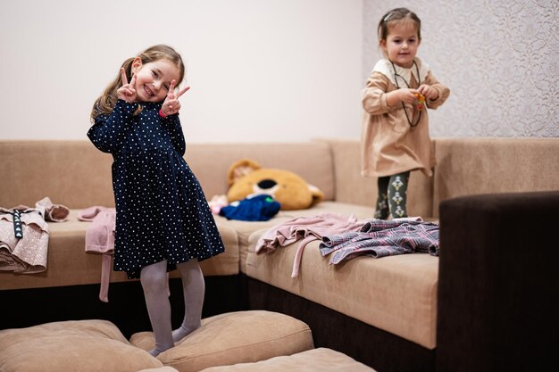 Zwei Schwestern wählen Kleidung aus dem Schrank zu Hause auf dem Sofa aus Mädchen zeigt eine Geste mit zwei Fingern